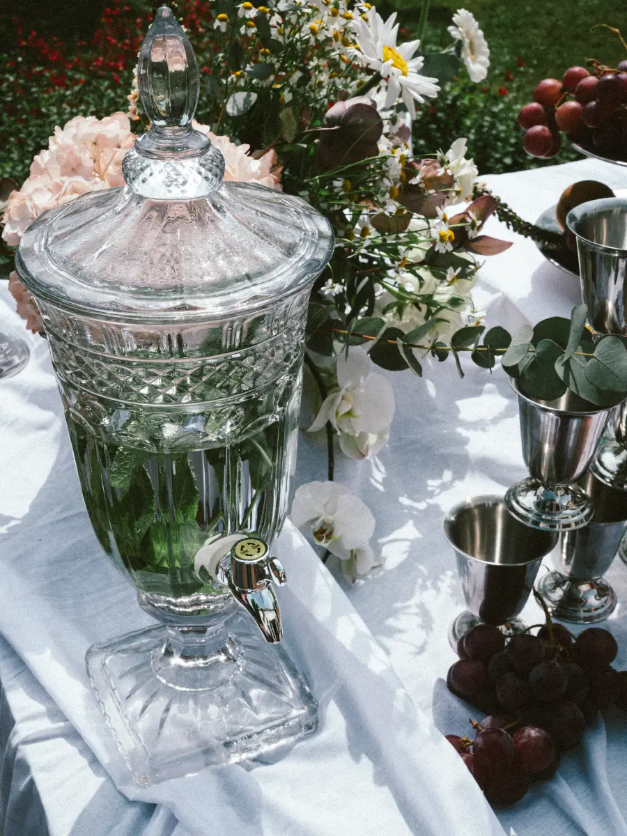 Fontaine à boissons argentée avec robinet, nappe blanche, fleur blanche et verre argenté sur table.