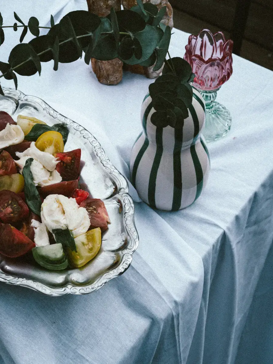 Salade tomate mozzarella fraîche pour vos événements (1) Salade, nappe blanche, vase blanc et noir avec tige de feuille, et verre.